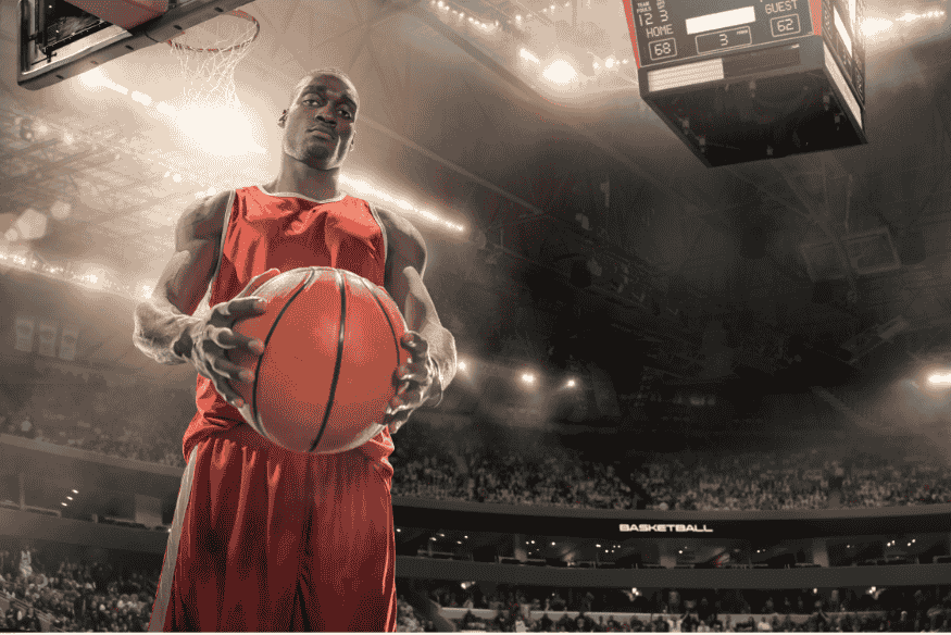 ogador de basquete segurando a bola com firmeza sob a cesta em ginásio iluminado.