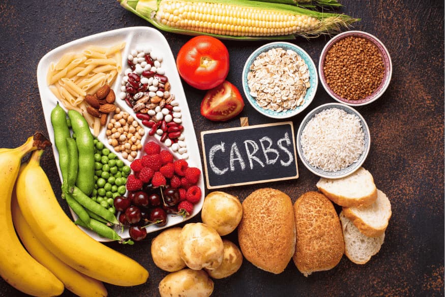 Alimentos fontes de carboidrato como pães, arroz, massas, frutas e cereais com placa escrita “carbs”.