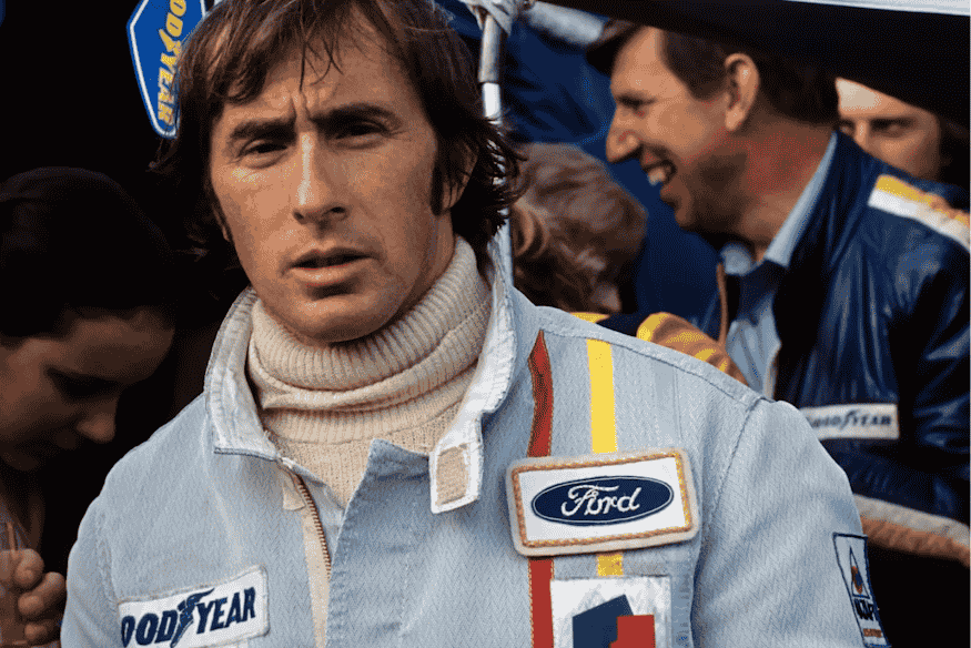 Piloto de Fórmula 1 vestindo macacão com logotipos da Ford e Goodyear, em meio a equipe de corrida.