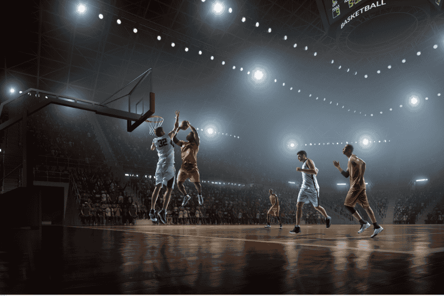 Jogadores em ação durante partida de basquete em ginásio com iluminação intensa.
