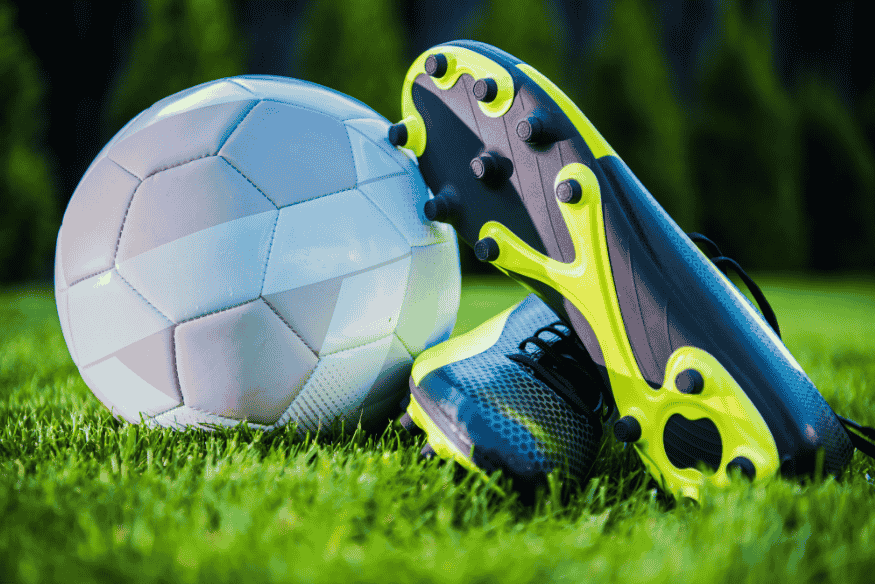 Chuteiras de futebol ao lado de uma bola em campo gramado, representando o início da competição.