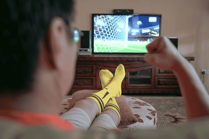 Pessoa assistindo a uma partida de futebol na TV usando meias de time, com o punho cerrado em comemoração.