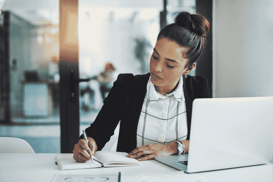 Mulher escrevendo anotações financeiras em caderno ao lado de um notebook, representando organização e disciplina após quitar dívidas para melhorar o score de crédito.